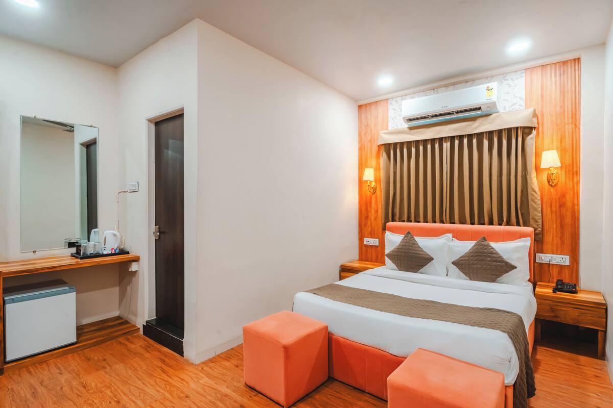 A hotel room with a bed, a desk, a chair, a mirror, a wardrobe, and an air conditioning unit.