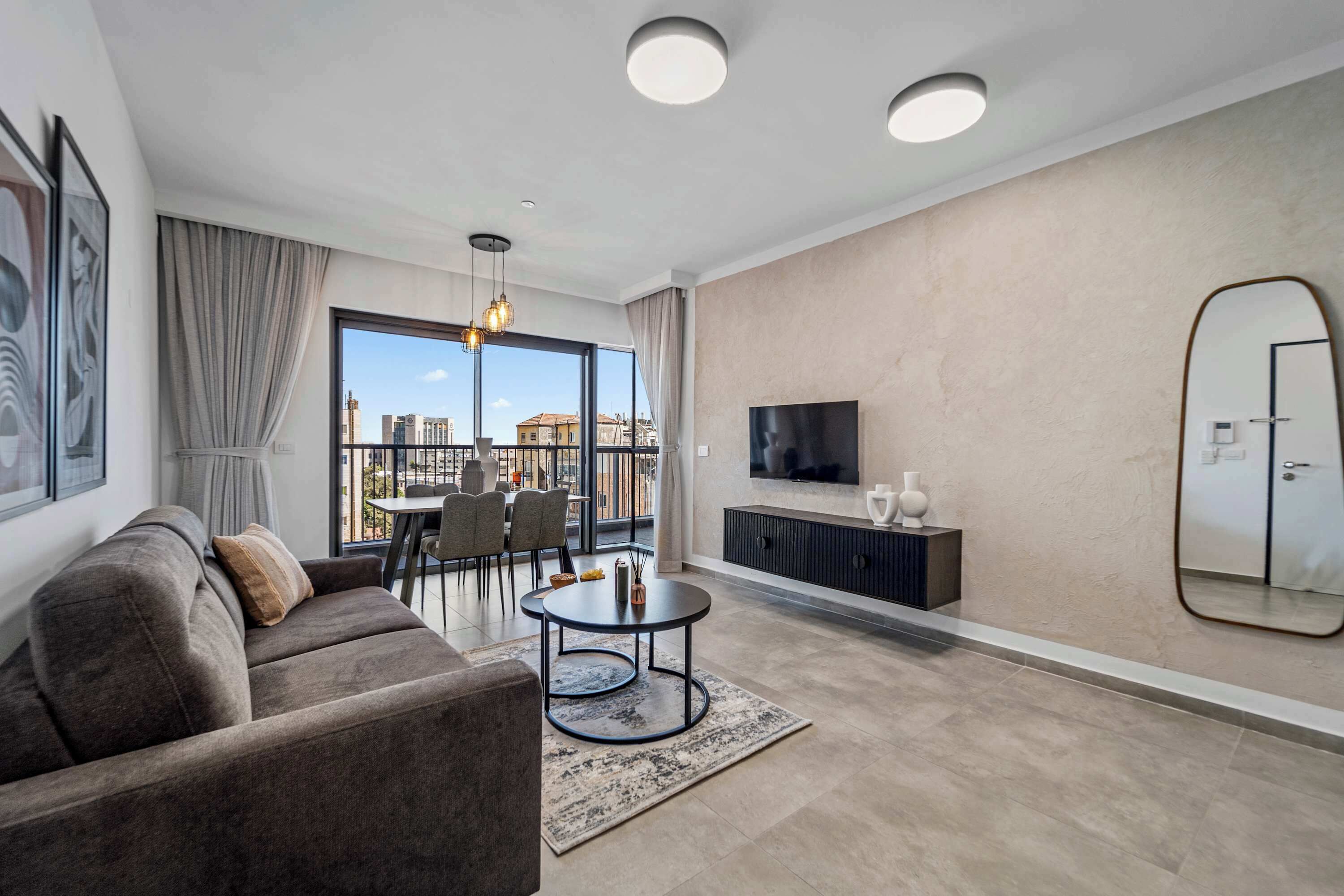 A modern living room with a sofa, a round coffee table, a TV, and a large window offering a city view.