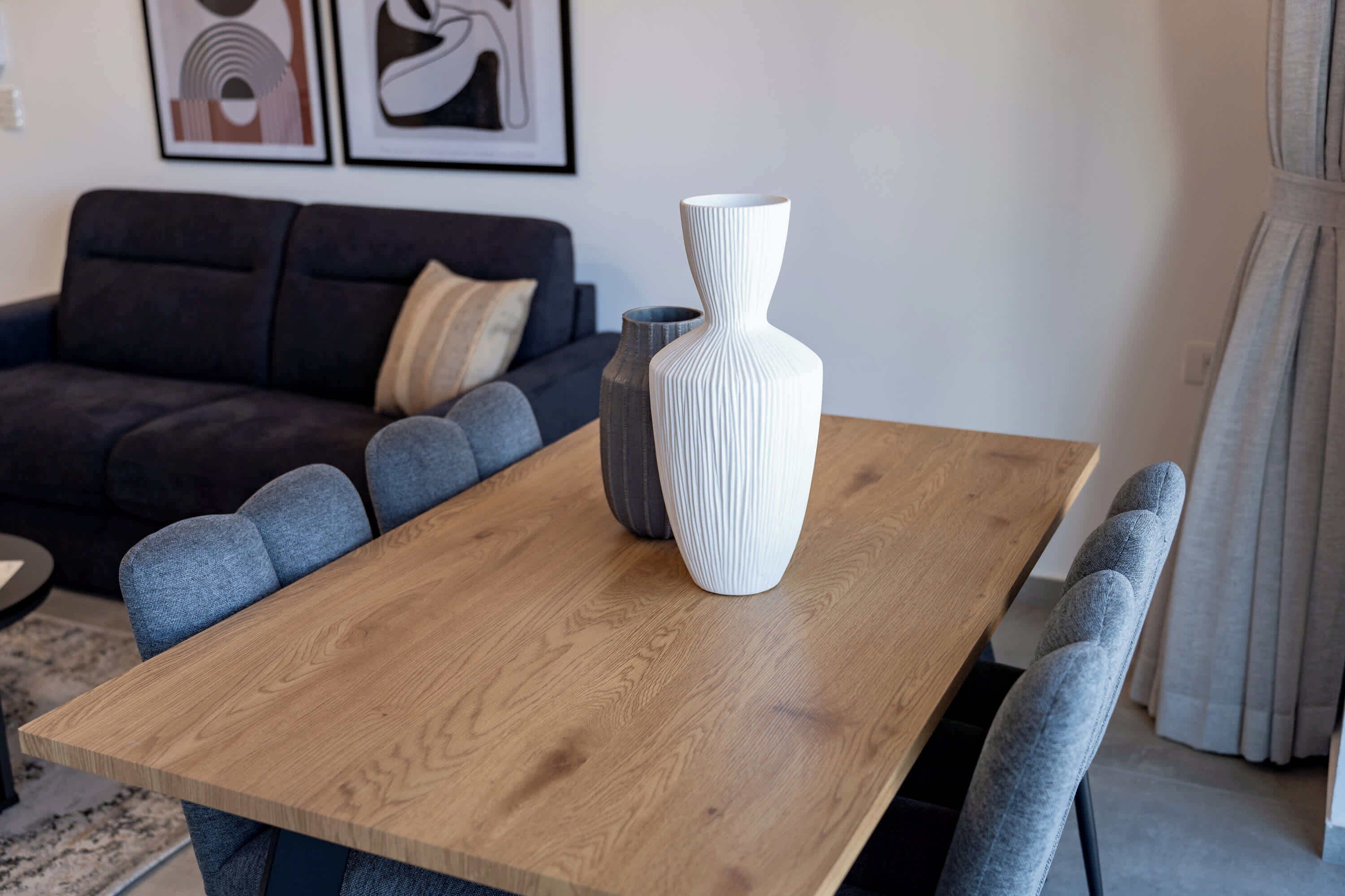 Ein Holztisch mit zwei Vasen, grau gepolsterte Stühle und eine dunkle Couch im Hintergrund.
