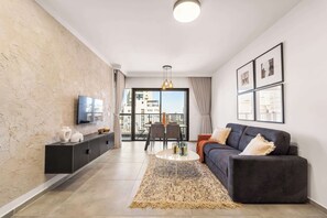 A modern living room with a sofa, a coffee table, and a TV mounted on the wall. There is a balcony visible through the glass door. - 1 Bedroom Savyon View - Isrentals (Jerusalem)