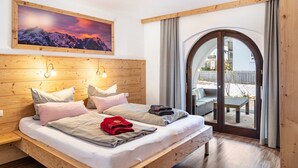 A bedroom with a wooden headboard, a large bed with pillows, a painting of mountains, and a view through a glass door to an outdoor seating area. - Apartment 'Bergwild' with Mountain View, Private Terrace and Wi-Fi (Garmisch-Partenkirchen)