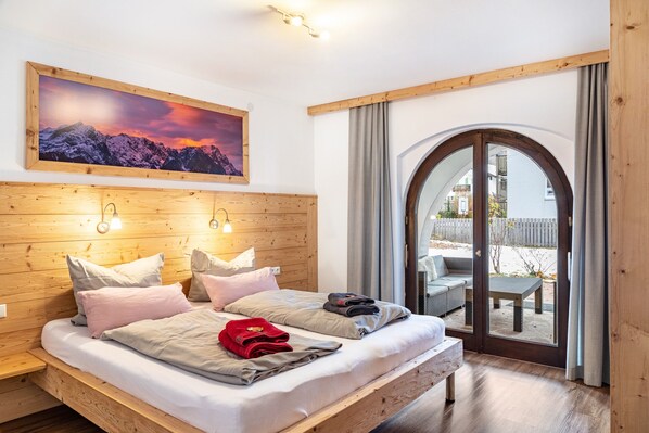 Un dormitorio con cabecera de madera, una cama grande con almohadas, un cuadro de montañas y vistas a una zona de estar exterior a través de un ventanal.