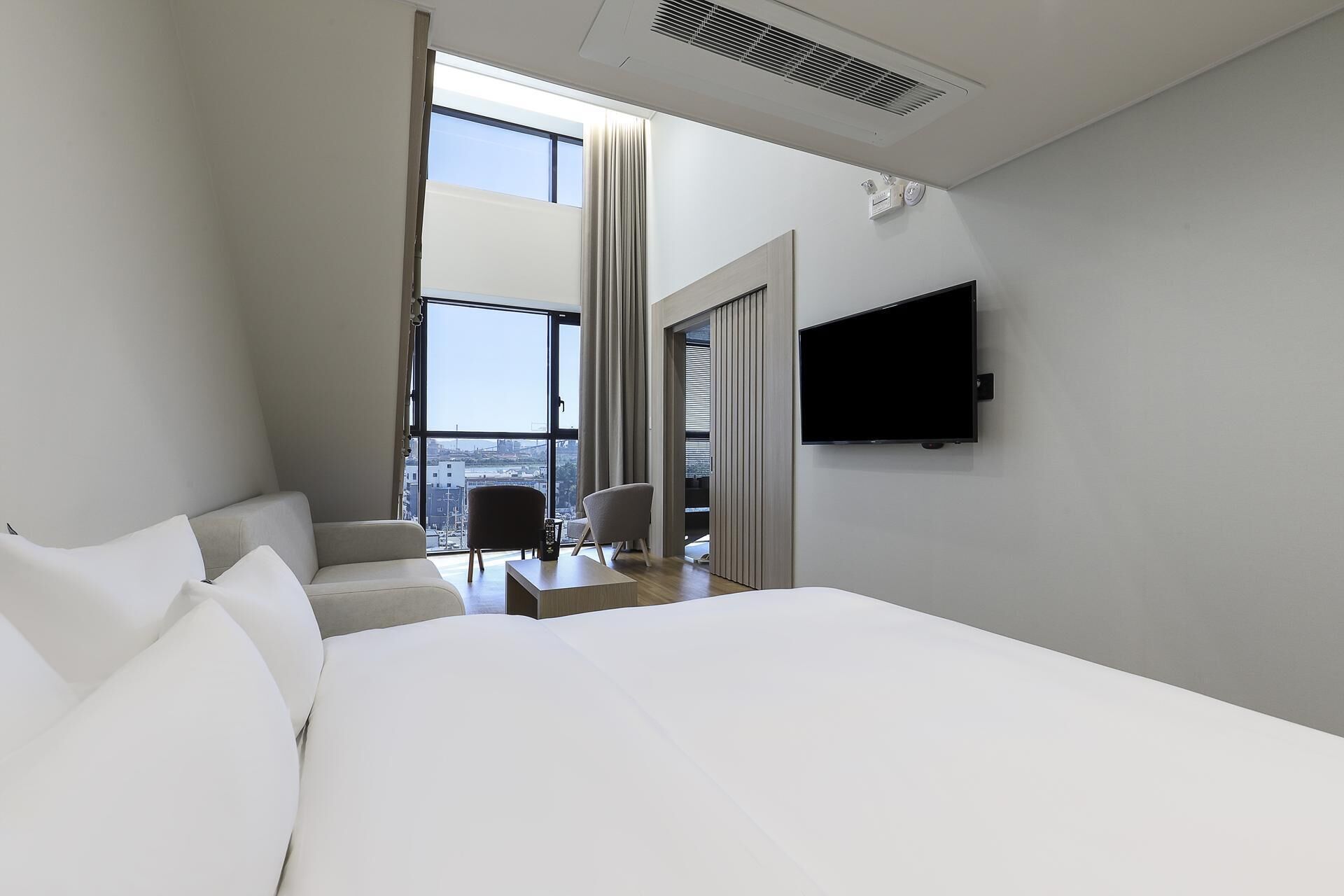 A modern hotel room with a large bed, a flat-screen TV mounted on the wall, and a view of the city through a large window.