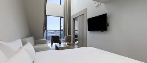 A modern hotel room with a large bed, a flat-screen TV mounted on the wall, and a view of the city through a large window.