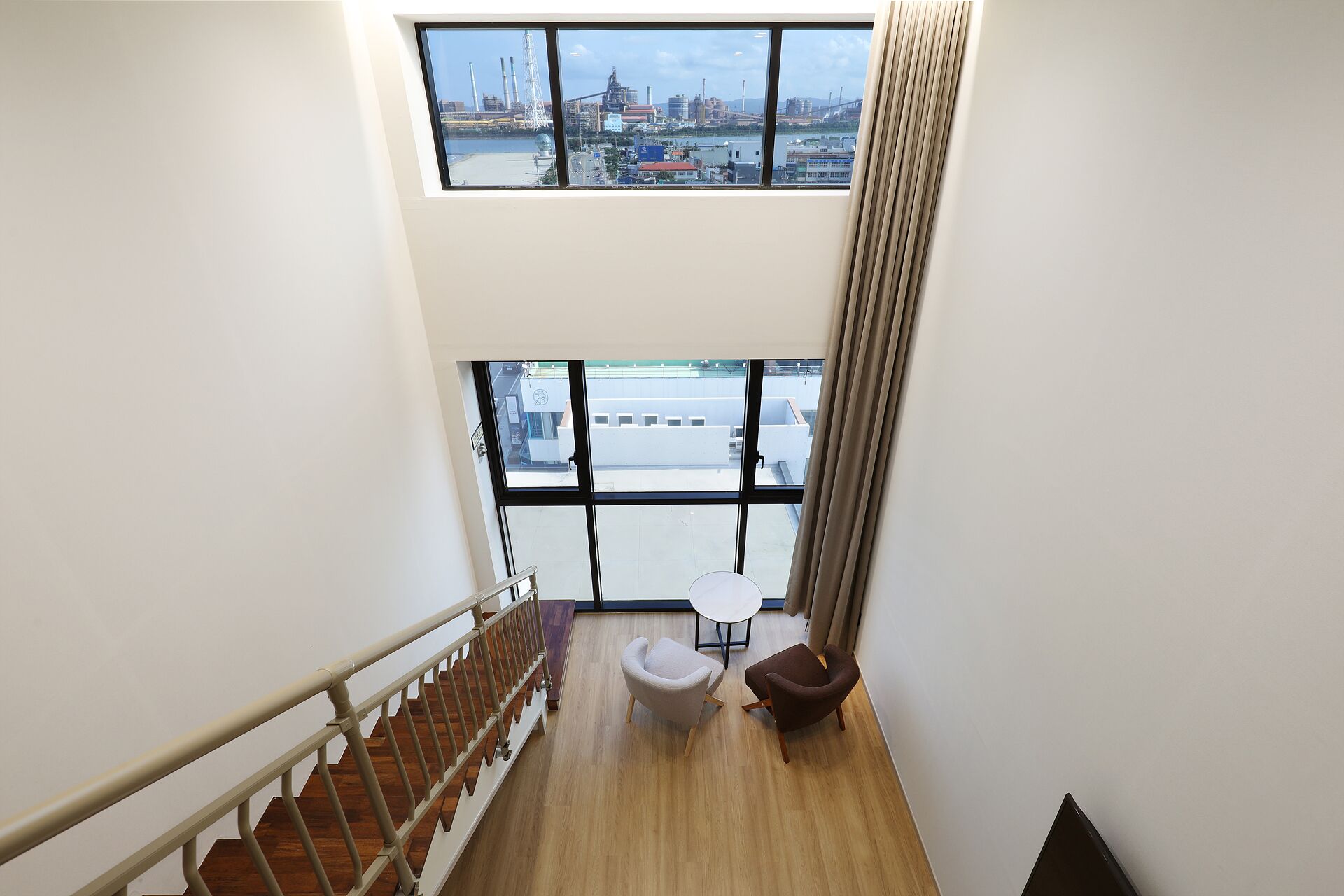 A modern interior with a wooden staircase, a seating area with two chairs and a small table, and large windows offering a view of an industrial area.