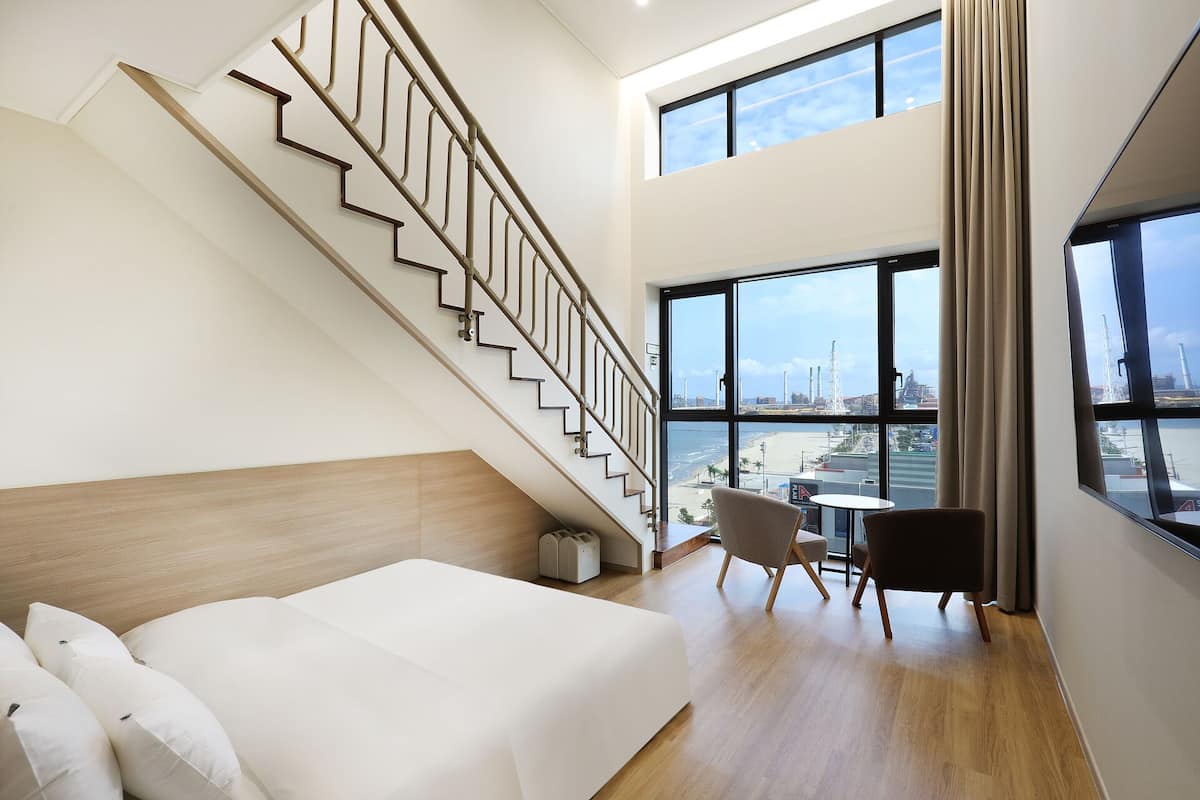 A modern hotel room with a wooden floor, a bed with white linens, a staircase, a view of the city through large windows, and a small table with a chair.