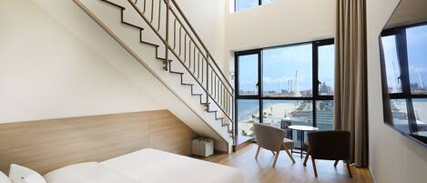 Quarto de hotel moderno com piso de madeira, cama com lençóis brancos, escada, vista da cidade através de grandes janelas e uma pequena mesa com uma cadeira.