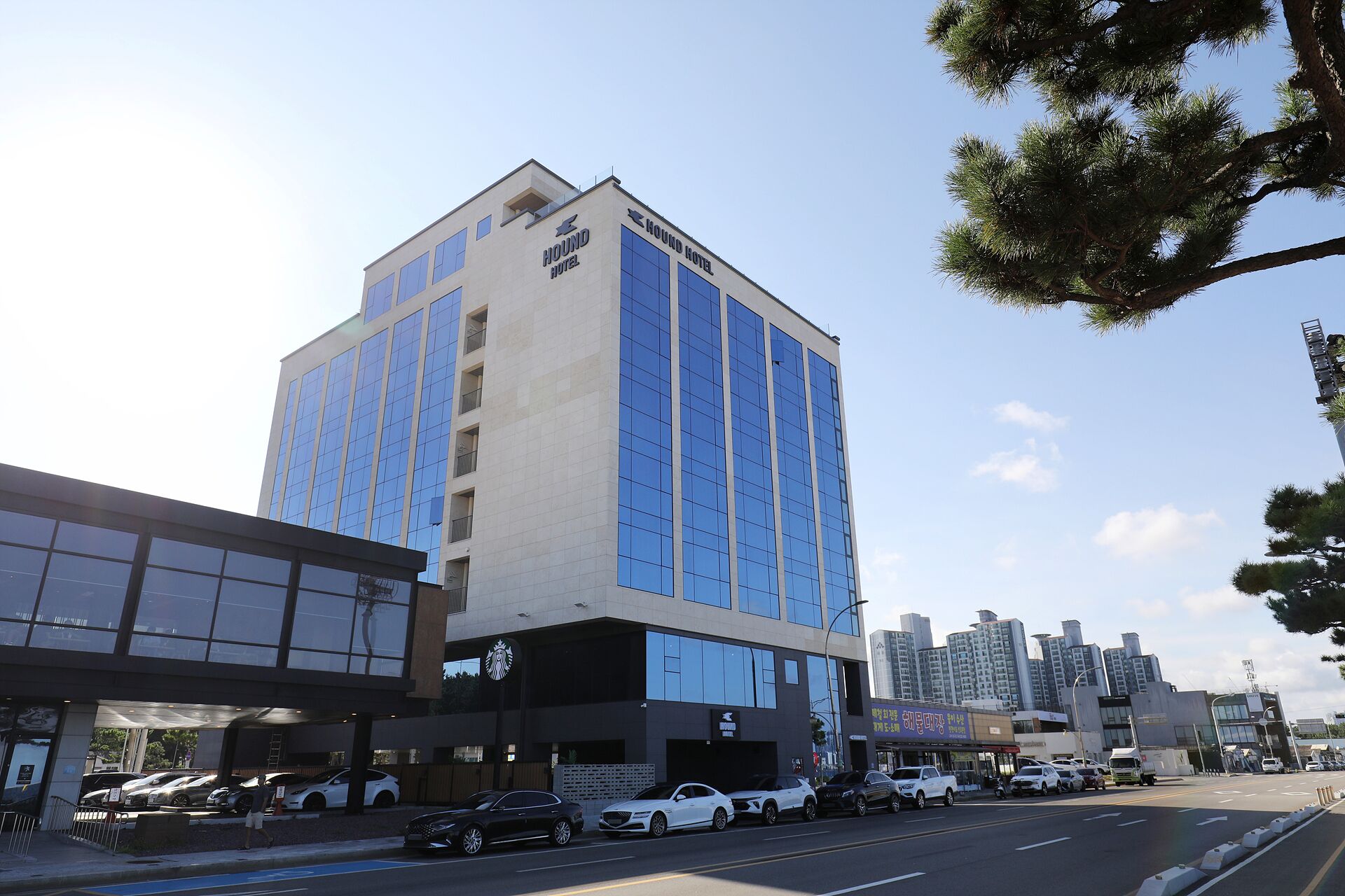 A modern building with a Hound Hotel sign, large windows, and a parking lot with cars.