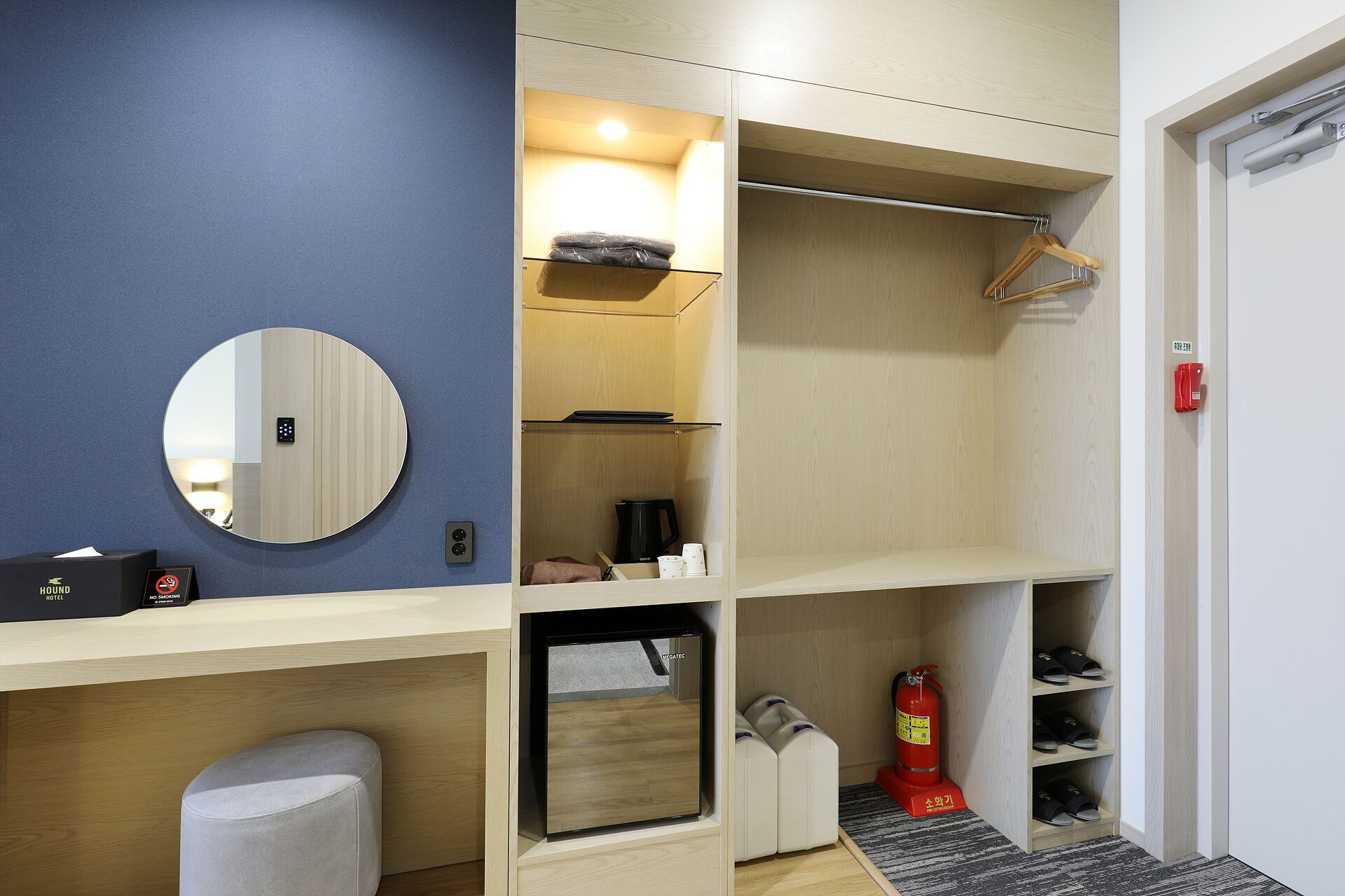 A hotel room with a blue accent wall, a round mirror, a minibar, a shoe rack, and a fire extinguisher.