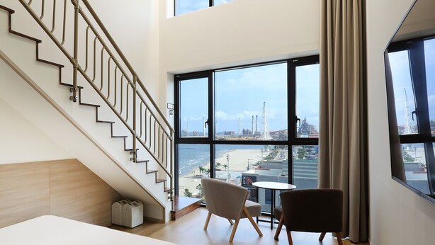 A modern hotel room with a staircase, a view of the beach and industrial structures, and a small round table with two chairs. - Pohang Hound Hotel Songdo Beach (Pohang)