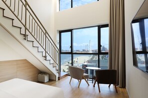 Una habitación de hotel moderna con una escalera, vistas a la playa y estructuras industriales, y una pequeña mesa redonda con dos sillas.