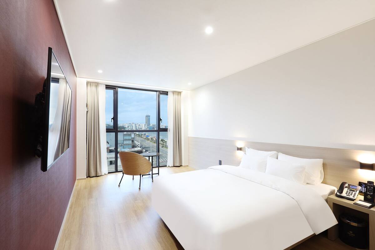 A hotel room with a large bed, a chair, a desk with a phone, and a view of a cityscape through large windows.