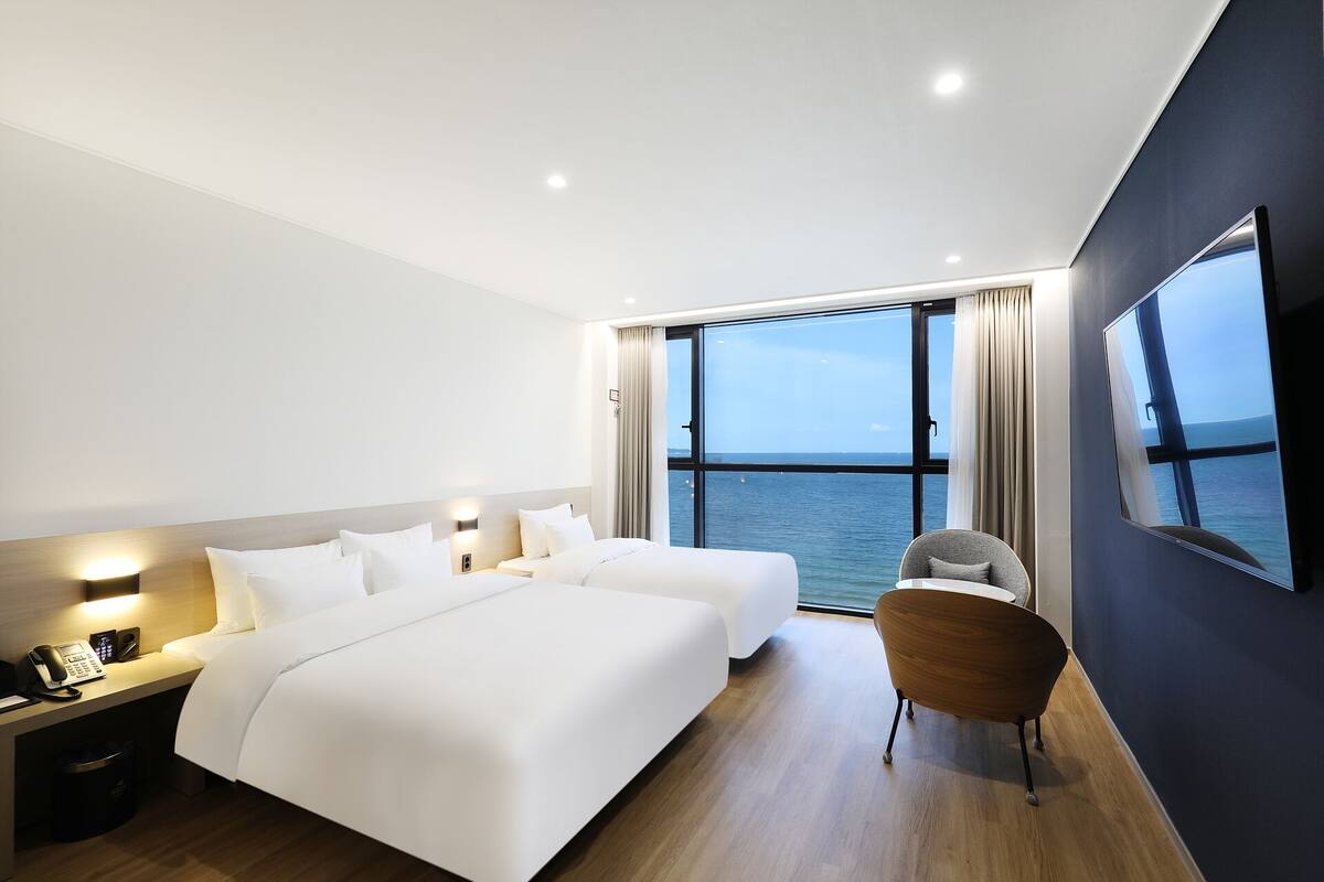A modern hotel room with two beds, a desk, a chair, and a large window offering a sea view.