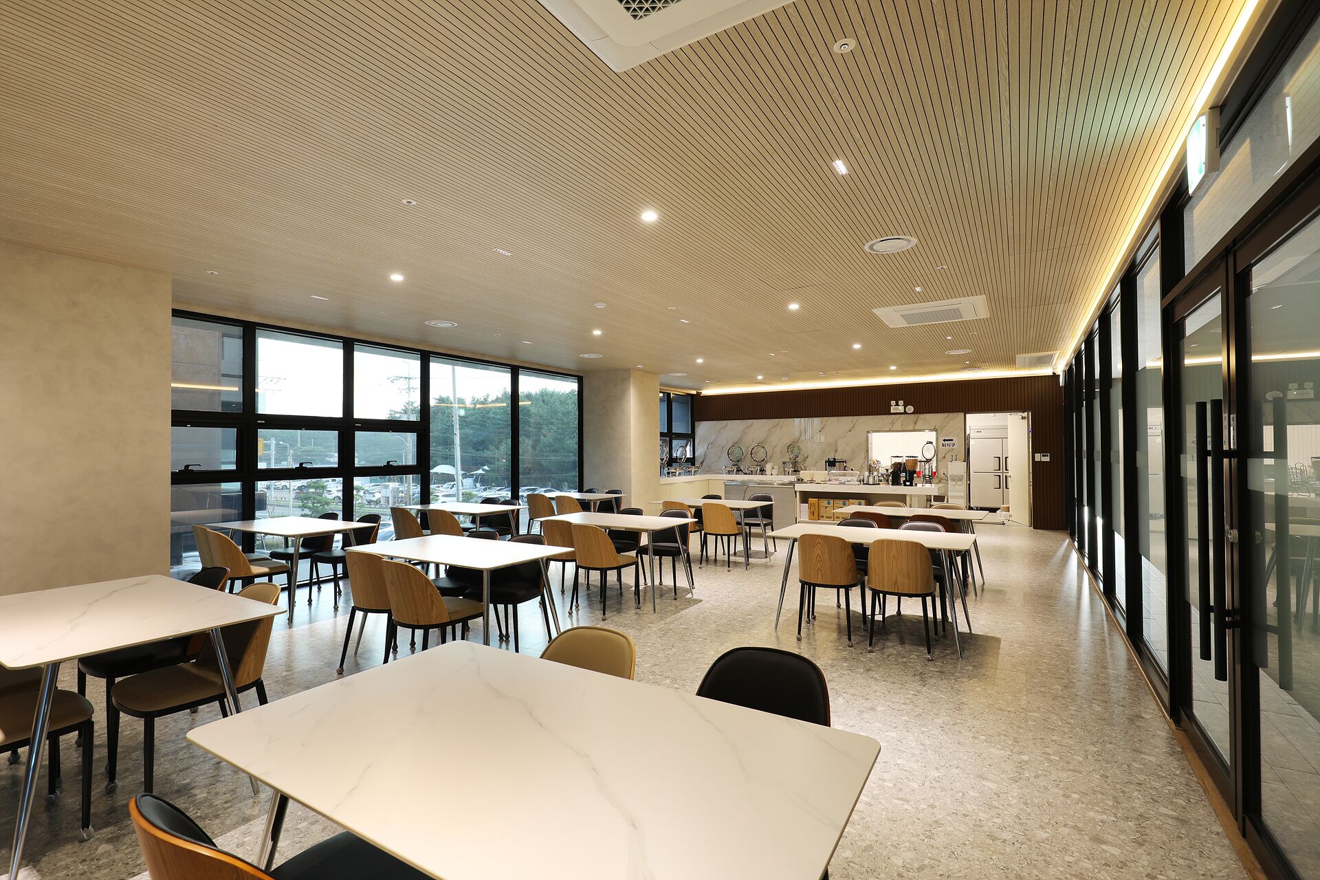 Un comedor moderno con mesas y sillas, ventanas amplias y una cocina al fondo.