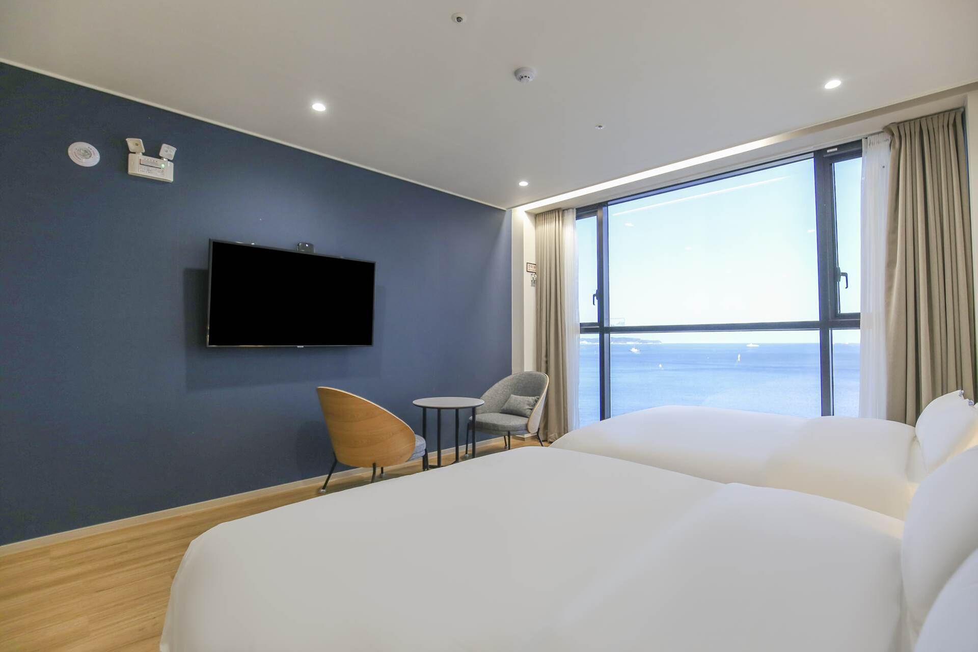 A hotel room with a large bed, a TV mounted on the wall, a small table, a chair, and a view of the sea through the window.