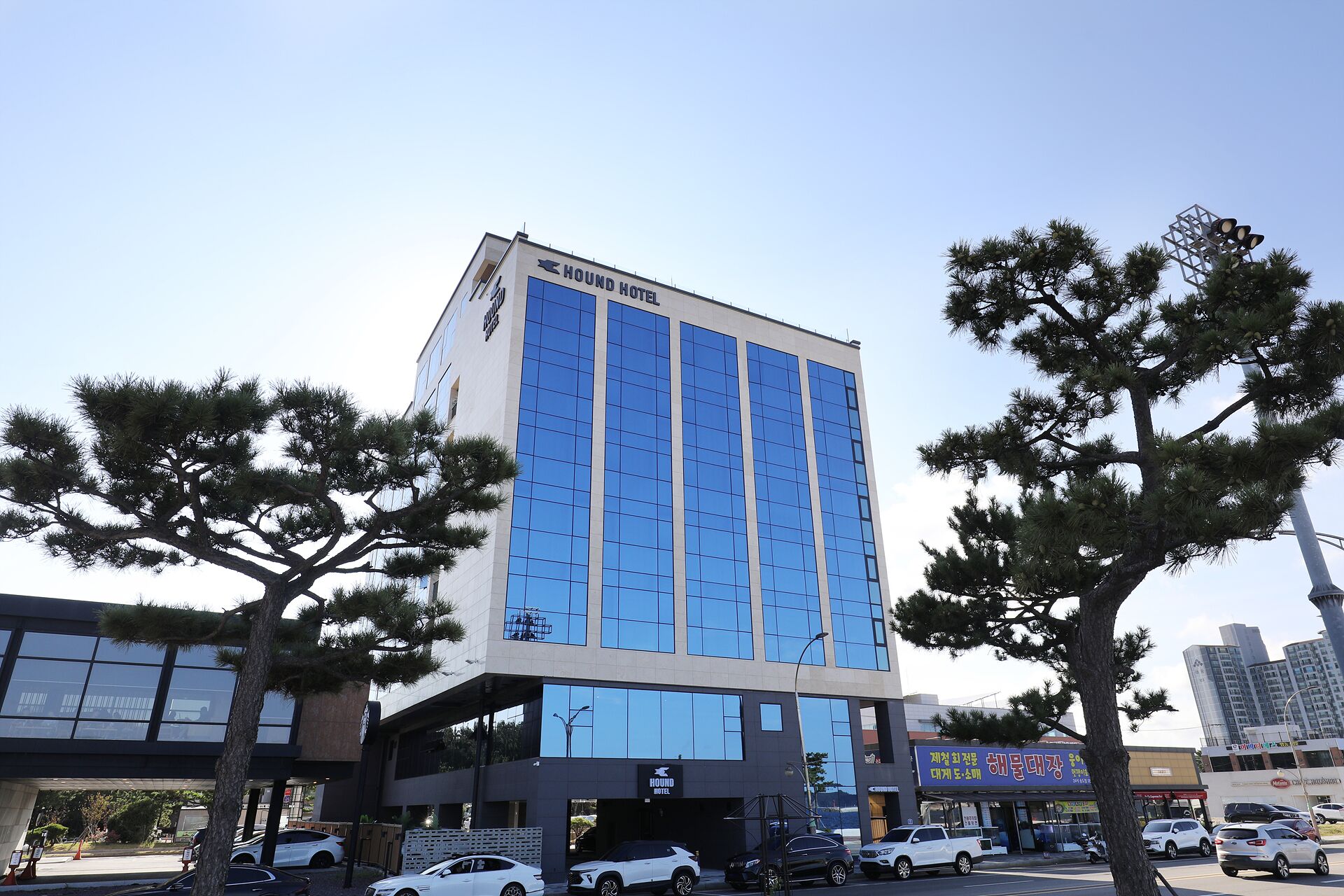 A modern hotel building with a prominent 'HOUND HOTEL' sign, large windows, and a parking area with several cars.
