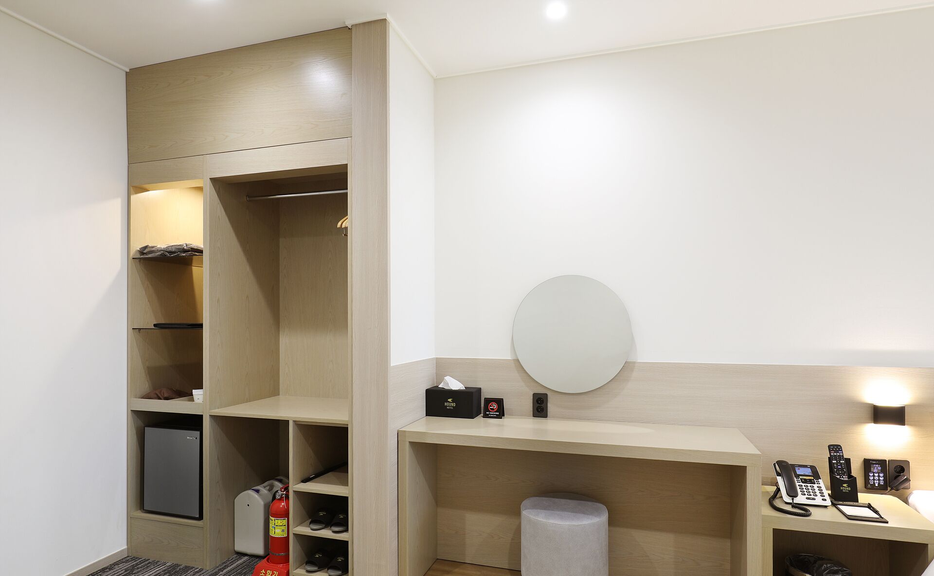 A hotel room with a wooden wardrobe, a small refrigerator, a fire extinguisher, a round mirror, a desk with a telephone, and a trash bin.