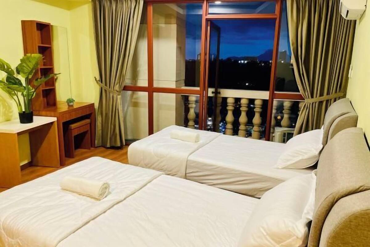 A hotel room with two beds, a desk, a small plant, and a view of the city at dusk.