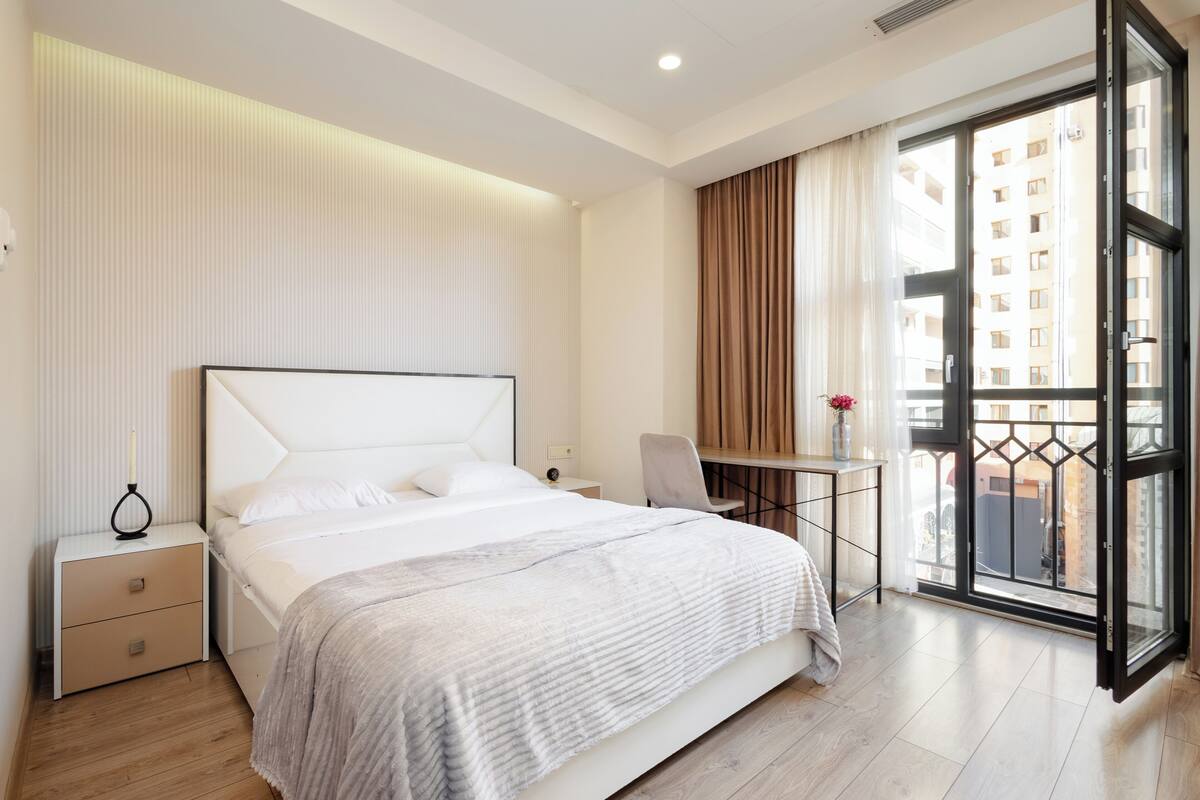 Ein modernes Hotelzimmer mit Bett, Nachttisch, Sessel, Schreibtisch und einem Balkon mit Blick auf Gebäude.