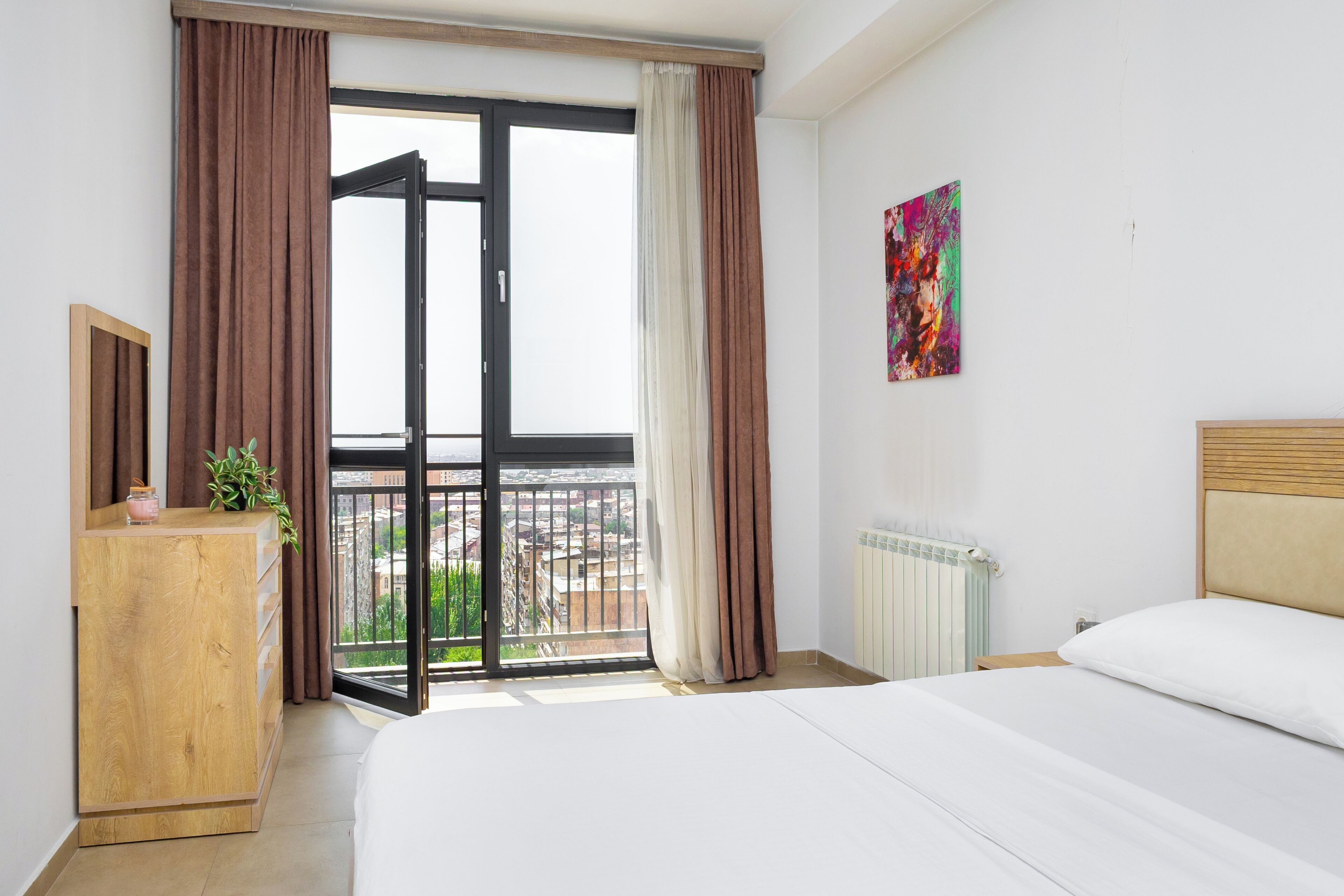 Quarto com cama, cómoda de madeira, radiador, janela com cortinas e vista para a cidade.