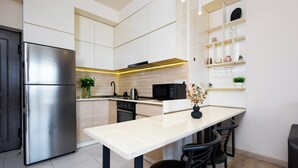 Una cocina moderna con un refrigerador de acero inoxidable, un microondas y un fregadero. Hay una encimera blanca y un comedor con sillas.