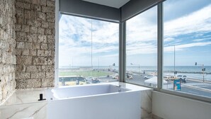 A modern bathroom with a large bathtub, a stone wall, and a view of a busy street and harbor through large windows. - w* | Scenic 3BR w/ Ocean View near C. Amurallada (Cartagena de Indias)