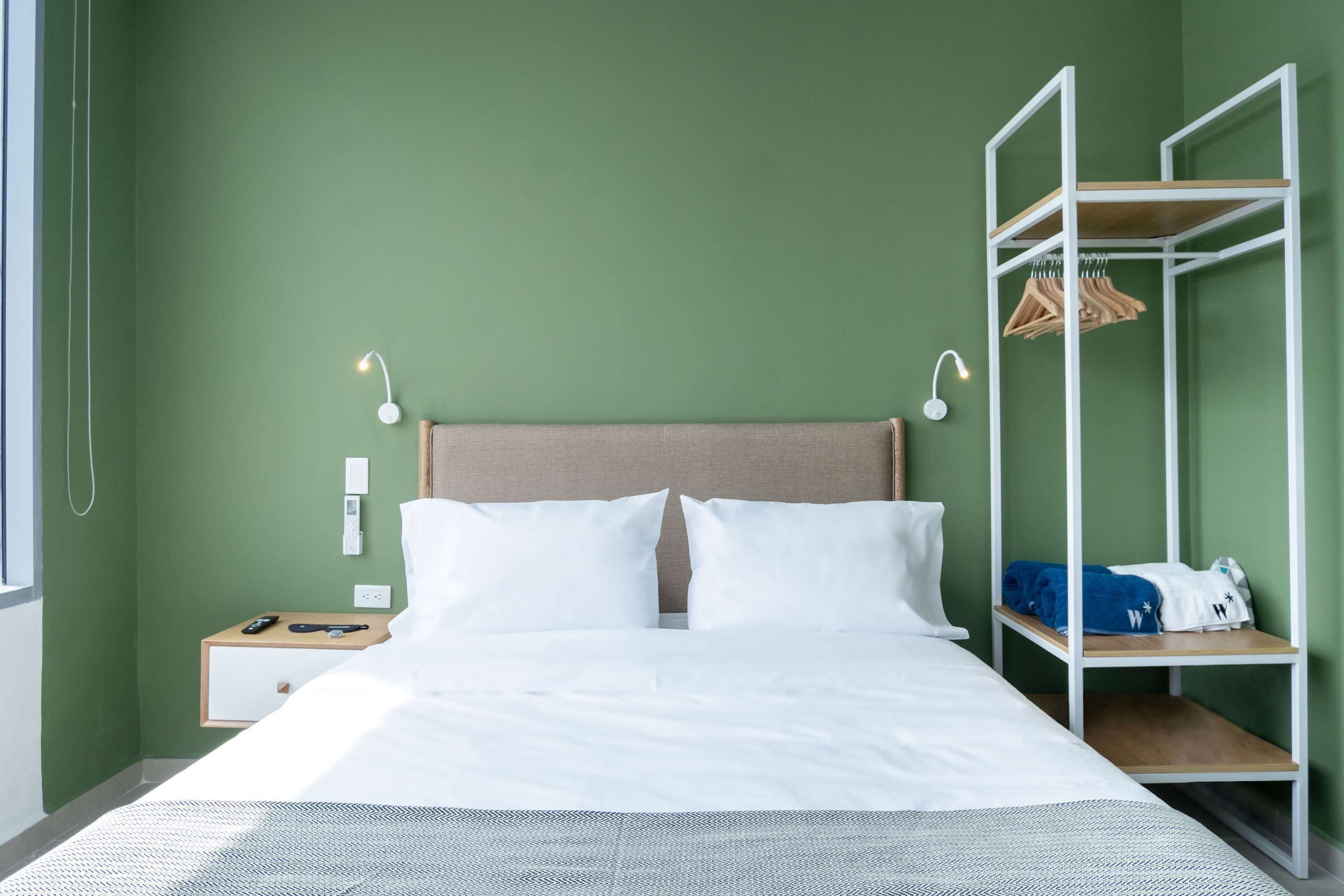 A neatly made bed with white linens, a bedside table with a lamp, a wooden nightstand, and a white wall with a green accent.