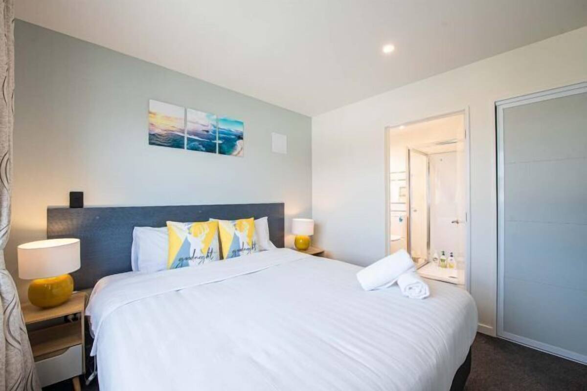 Une chambre d’hôtel avec un grand lit, une table de chevet avec une lampe, une œuvre d’art accrochée au mur et une porte donnant sur une salle de bain.
