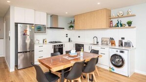 Eine moderne Küche mit weißen Schränken, einem Edelstahlkühlschrank, einem Holzessttisch mit Stühlen, einem Einbauofen und einer Waschmaschine.