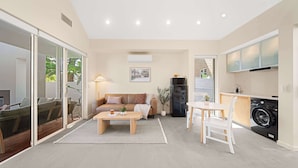 Ein modernes Wohnzimmer mit einer Couch, einem Couchtisch und einem Essbereich mit Stühlen. Es gibt eine Glasschiebetür, die auf einen Balkon führt, sowie eine Waschmaschine und ein Kühlschrank im Küchenbereich.