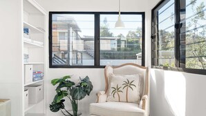 A room with a large window, a chair with a palm tree pillow, a potted plant, and a white shelving unit. - Moffat Beach Abode - Dog-friendly Escape by the Sea (Caloundra Region)