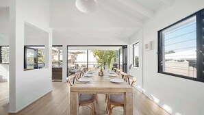 A dining area with a long table, chairs, and a view through large windows. - Moffat Beach Abode - Dog-friendly Escape by the Sea (Caloundra Region)