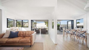 A modern living room with a sofa, coffee table, and dining area with chairs and a table. - Moffat Beach Abode - Dog-friendly Escape by the Sea (Caloundra Region)