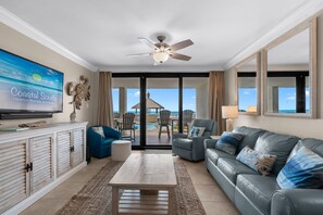 A living room with a television, a ceiling fan, a sofa, a coffee table, and a view of the ocean through large glass doors. - Beachfront 3 Bed Ground Floor at Summer House in OB! Free Daily Activities! (Orange Beach)