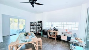 A spacious room with a ceiling fan, a large window with a view of trees, a wooden table with a glass top, and a kitchenette area.