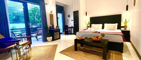 Una habitación de hotel moderna con una cama grande, una mesa de comedor con fruta, un rincón de estar con sillas y vistas al exterior.