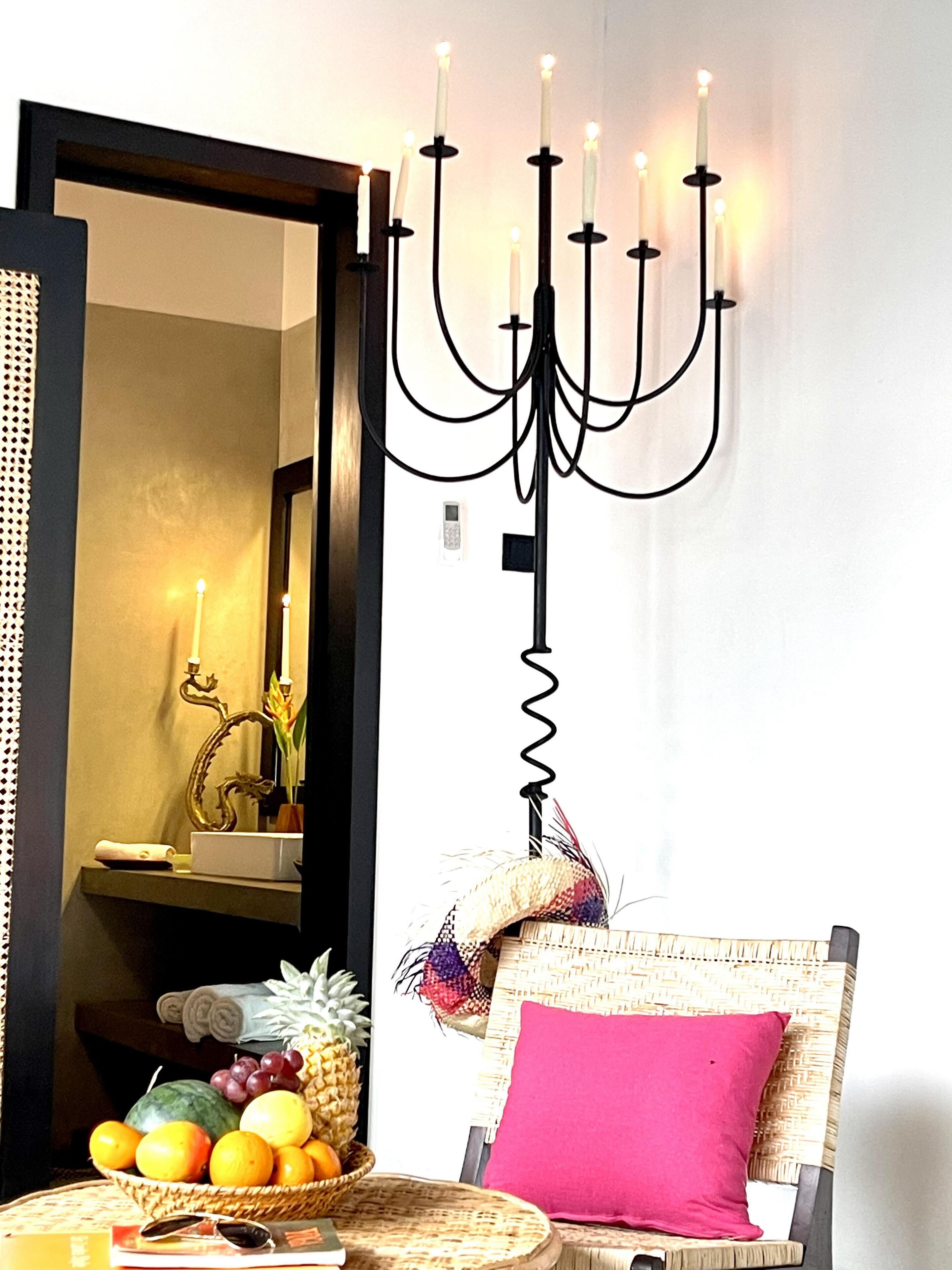 Une pièce avec un miroir, un fauteuil en osier agrémenté d’un coussin rose, un panier de fruits et un chandelier fixé au mur.