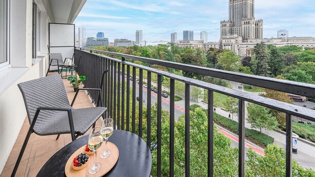 A balcony with a table set for two, a potted plant, and a view of a cityscape including a prominent building. - City center Apart near Palace of Culture (Warsaw)