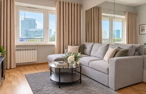Un salon moderne avec un canapé gris, une table basse en verre et une vue sur les immeubles de la ville, visible à travers de grandes fenêtres.