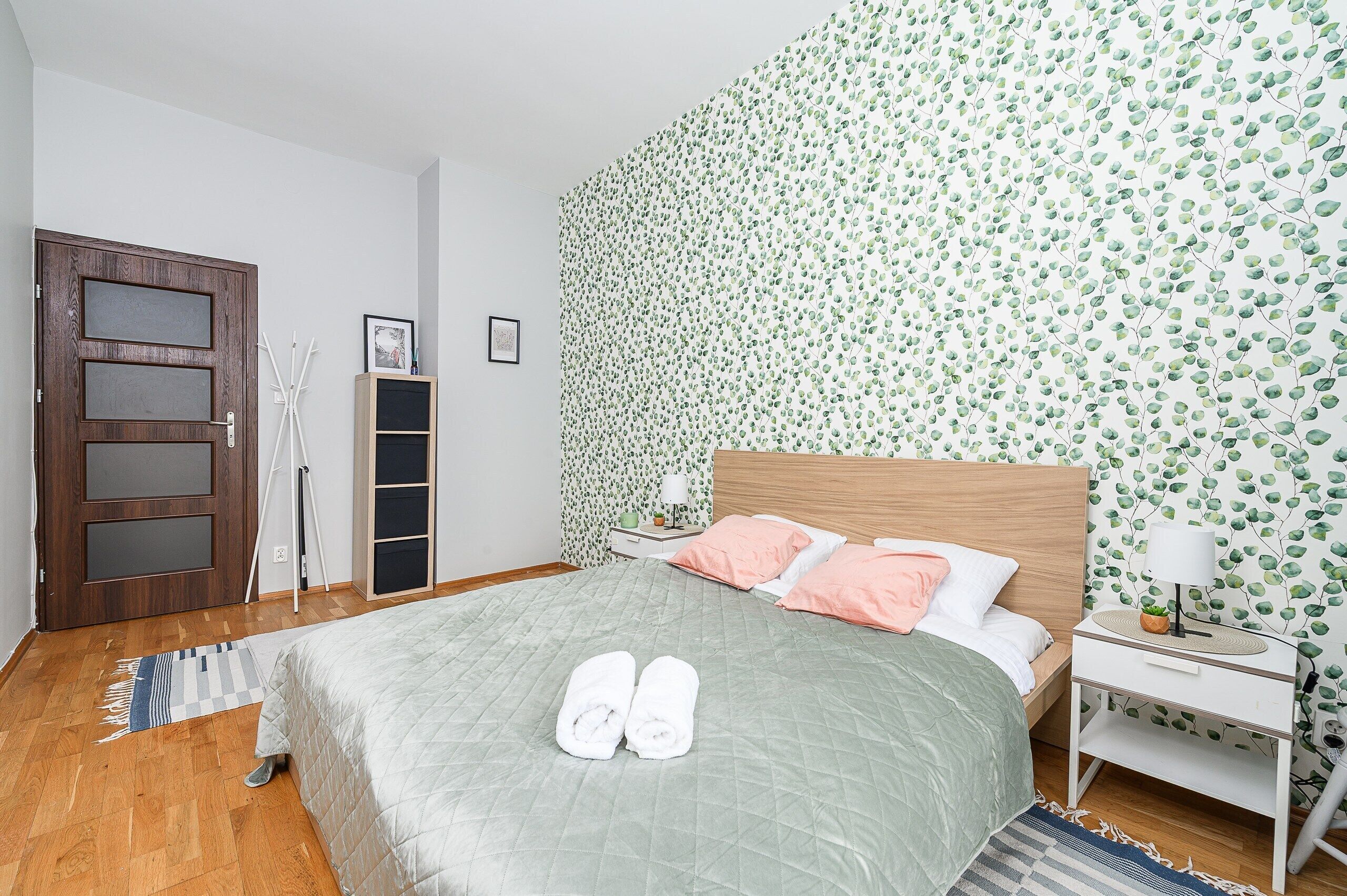 A bedroom with a bed, a nightstand, a wooden door, and a wall with a leafy pattern.