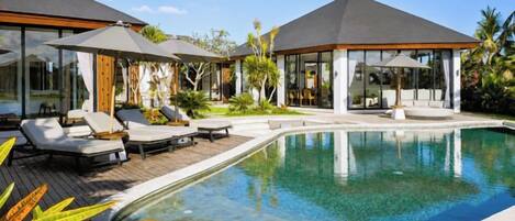 A modern pool area with lounge chairs and umbrellas, surrounded by tropical plants and a clear blue sky.