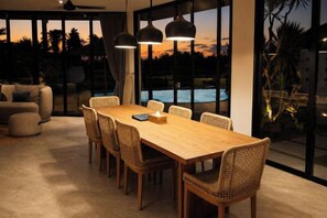 A dining area with a wooden table and wicker chairs, large windows with a view of a swimming pool, and a ceiling fan. - Villa Vidrev - Private Infinity Pool & Rice Fields by BaliSuperHost (Bali)