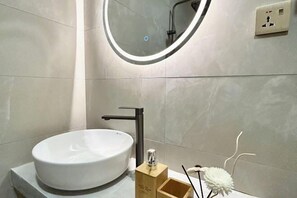 Un baño moderno con un lavabo blanco redondo, un grifo negro, un dispensador de jabón de madera y un espejo con luz circular.