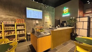 A food counter with a menu board, a fruit display, and seating area with yellow chairs. - ONE18 Boutique Hotel (Kuala Lumpur)