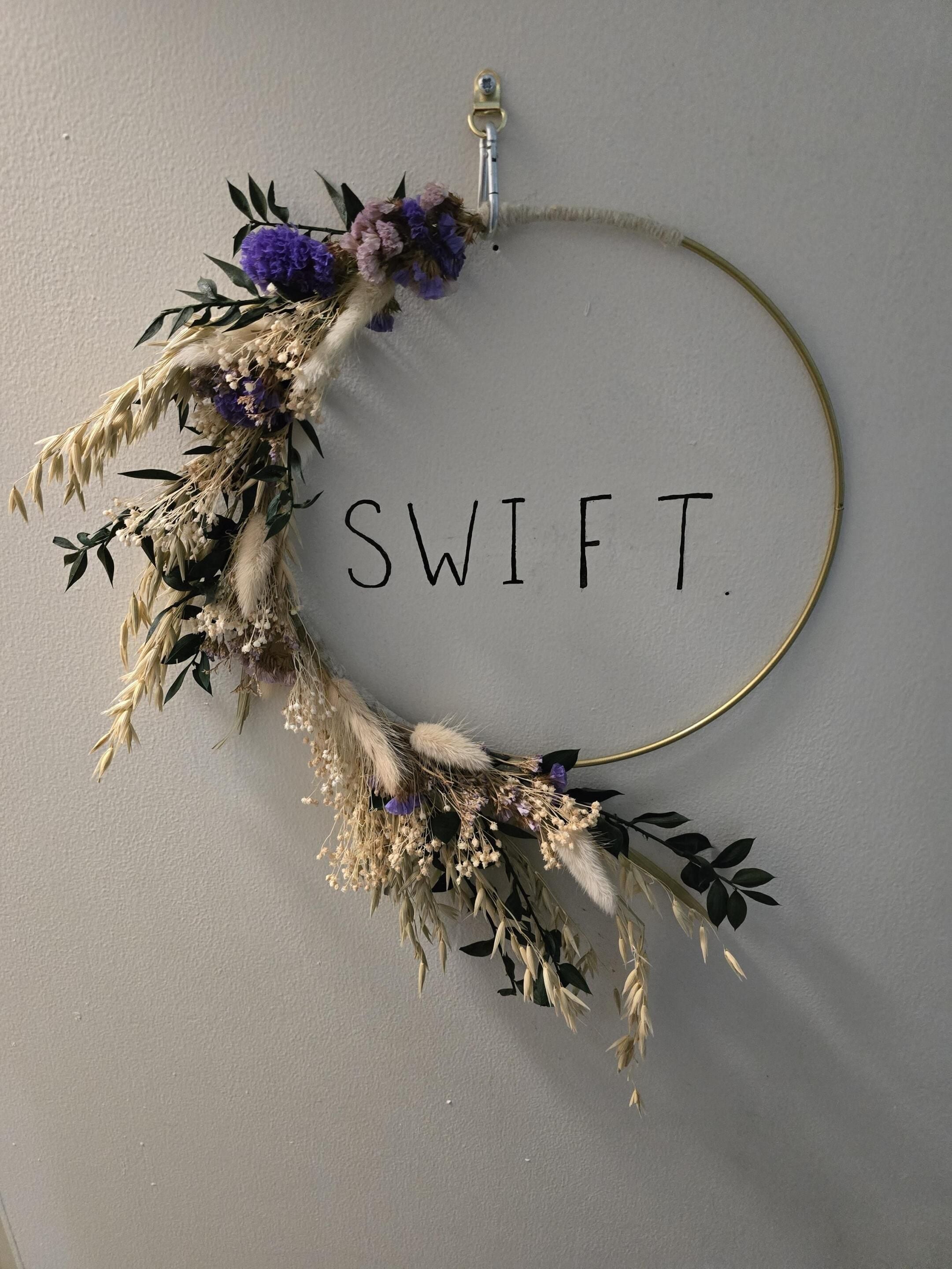 Ein an der Wand befestigter dekorativer Kranz mit dem Wort 'SWIFT' und einem goldfarbenen, runden Rahmen.