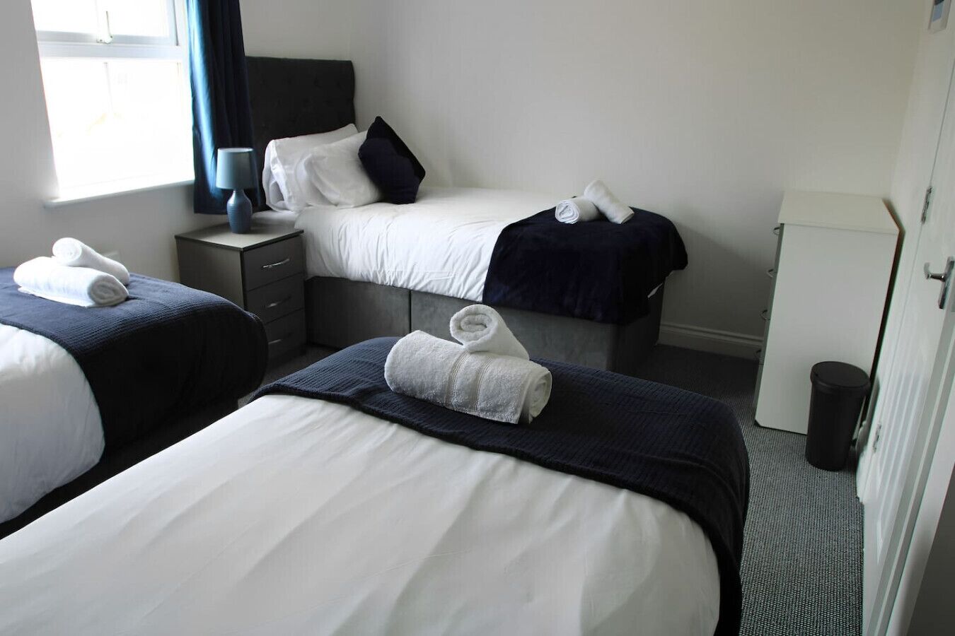 Ein Hotelzimmer mit zwei Betten, jedes mit weißen Laken und dunklen Kopfteilen. Auf den Betten liegen Handtücher und ein kleiner Tisch mit einer Lampe.