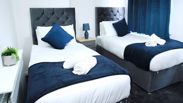 Habitación de hotel con dos camas, cada una con cabecero oscuro y ropa de cama blanca. Hay dos mesitas de noche, una con una lámpara y la otra con una planta. La habitación tiene un diseño moderno y obras de arte en las paredes.