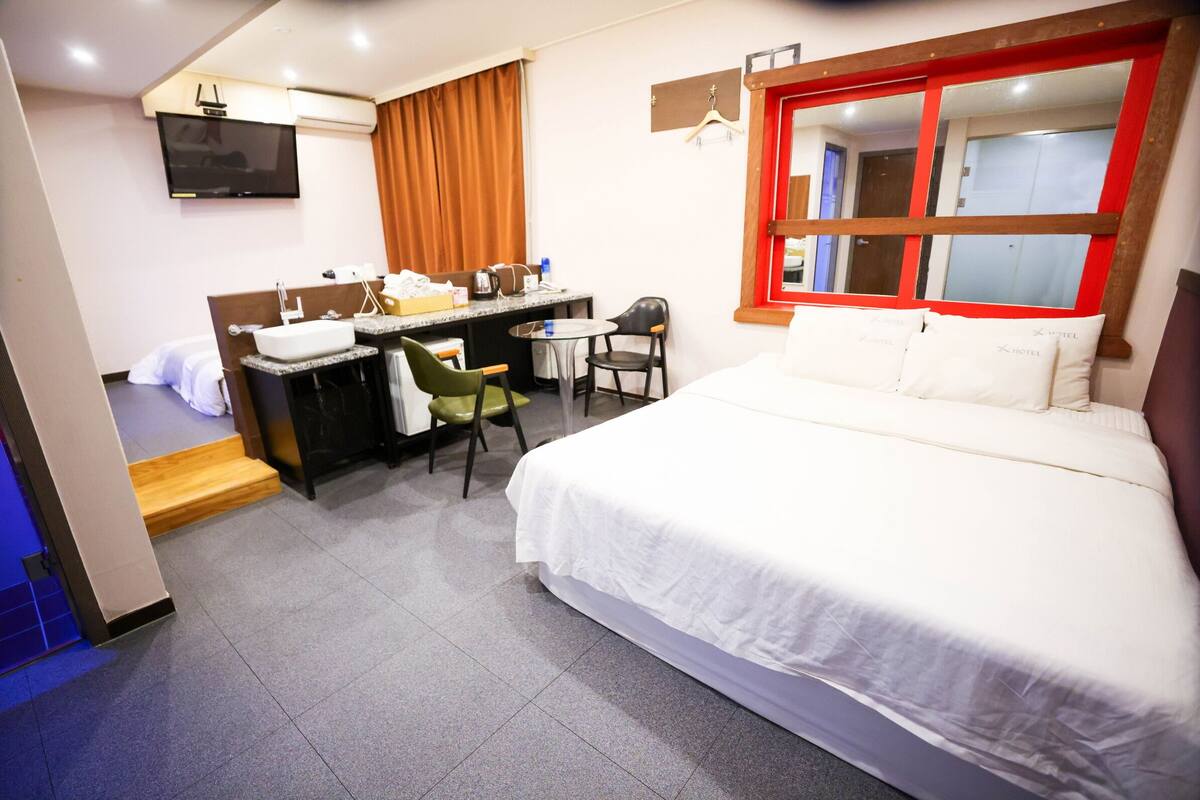 A hotel room with a bed, a desk with a sink, a chair, a TV mounted on the wall, and a window with a red frame.