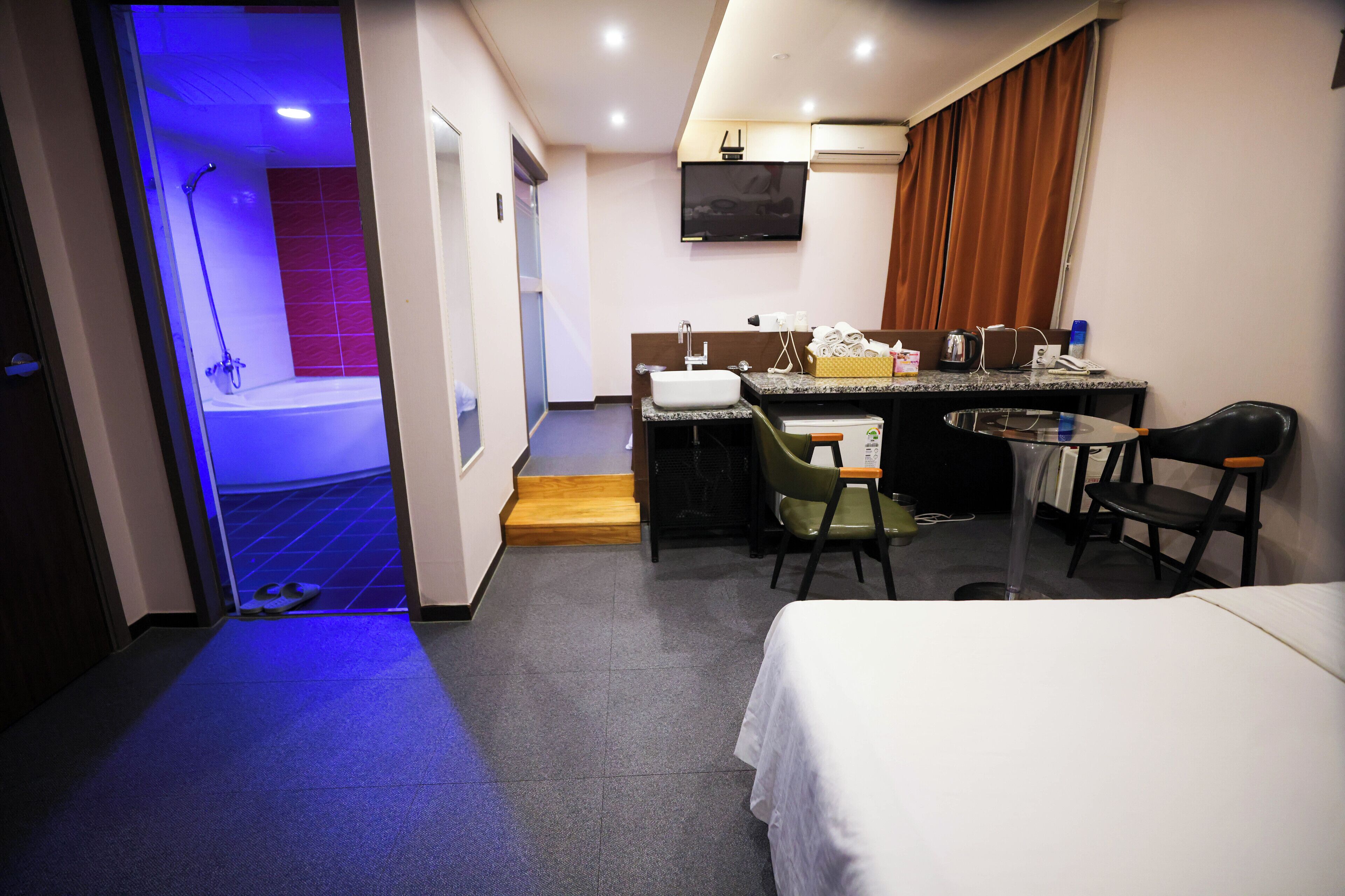 A hotel room with a bed, a small desk with a chair, a sink, and a shower area.