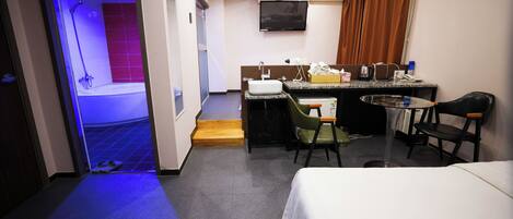 A hotel room with a bed, a small desk with a chair, a sink, and a shower area.